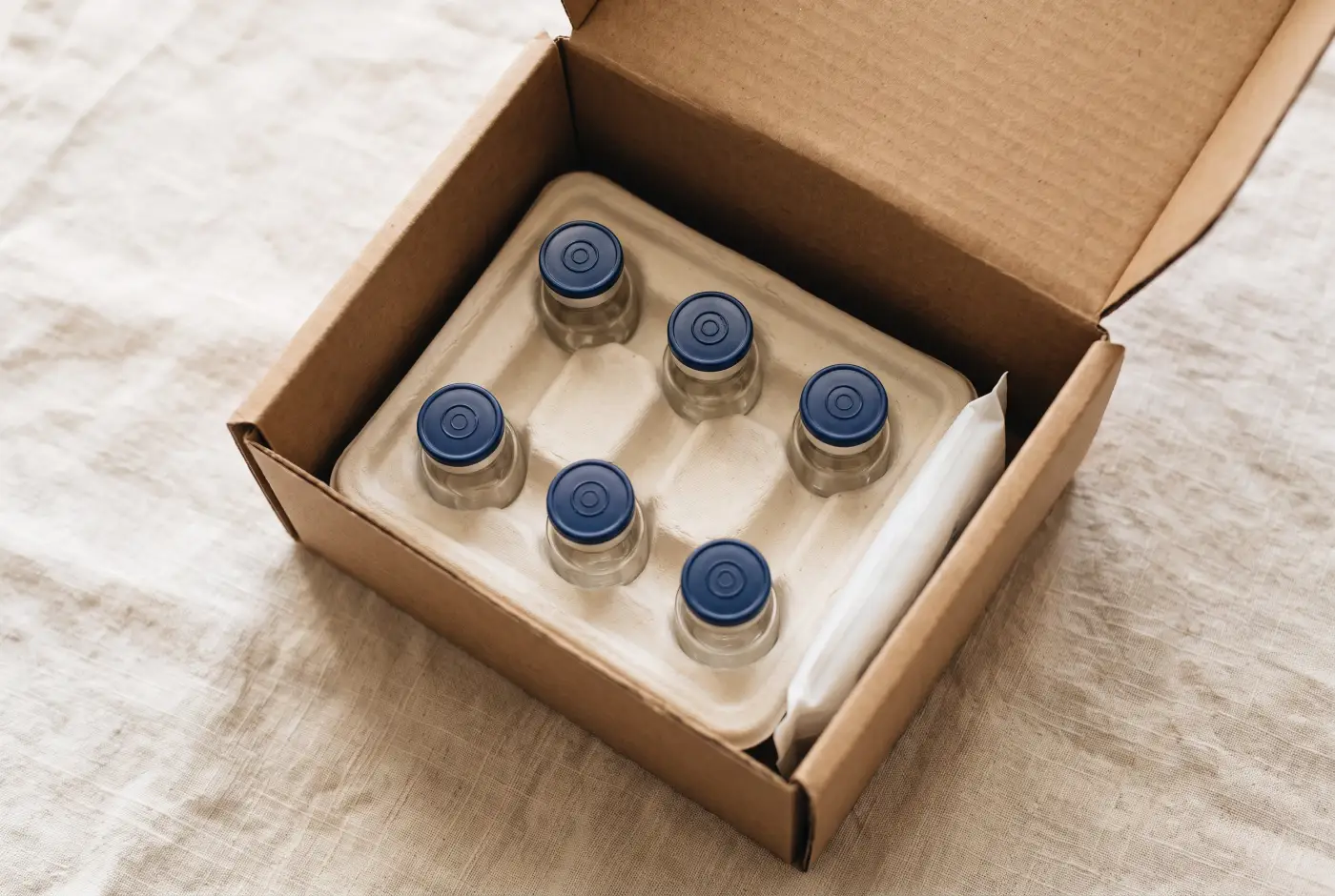 An open shipping box with navy-capped glass vials nested in a paper insert beside an ice pack