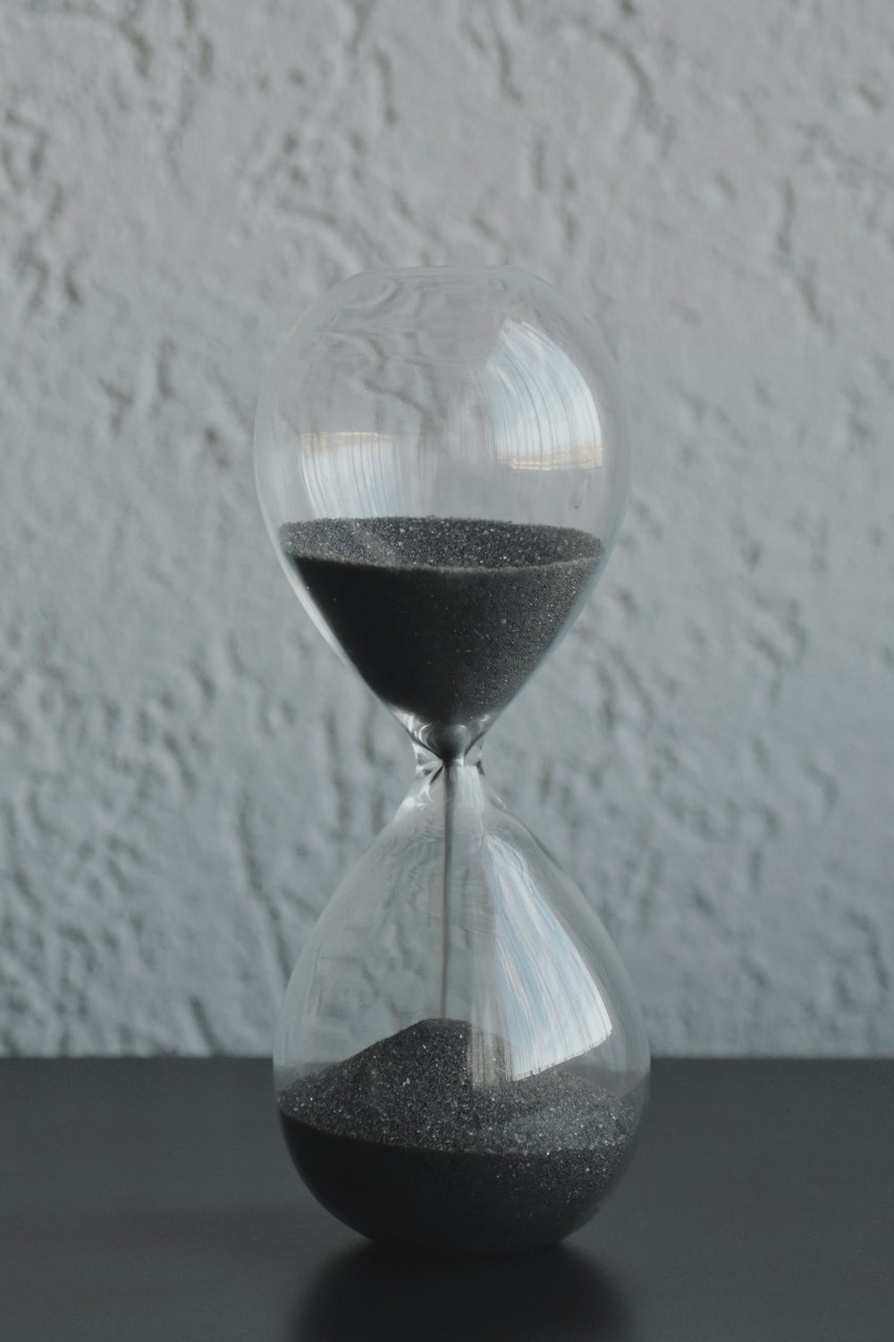 A glass hourglass with dark sand running, on a dark tabletop against a pale textured wall