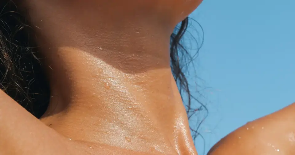 Close-up of a woman's sunlit face, neck, and shoulders with dewy, healthy-looking skin against a clear blue sky.