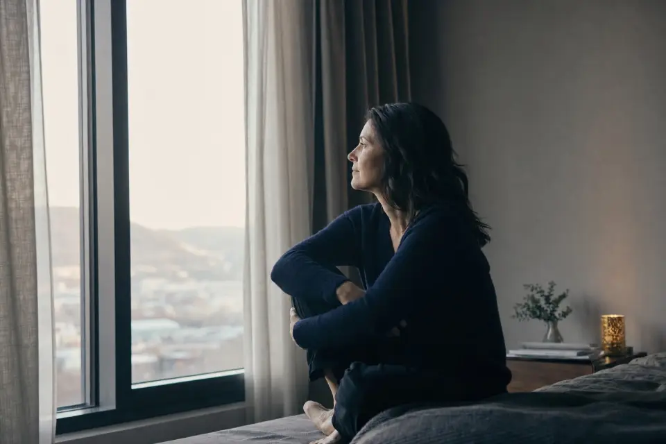 A woman sitting by a tall window in a quiet room, looking out at overcast daylight