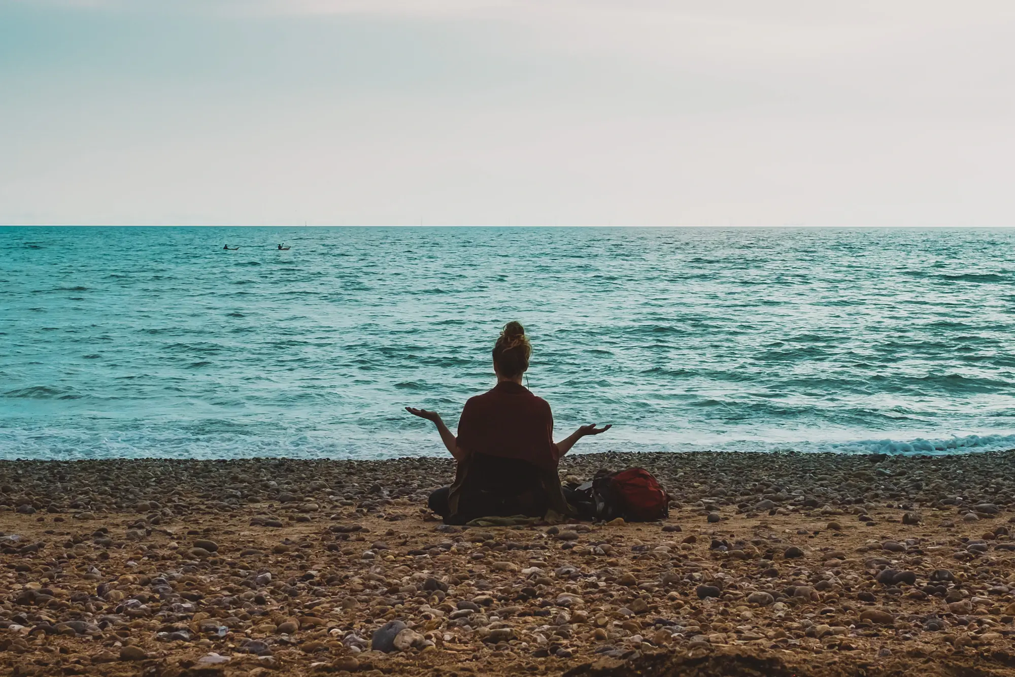 A woman sitting calmly on a pebble beach facing the ocean at dawn