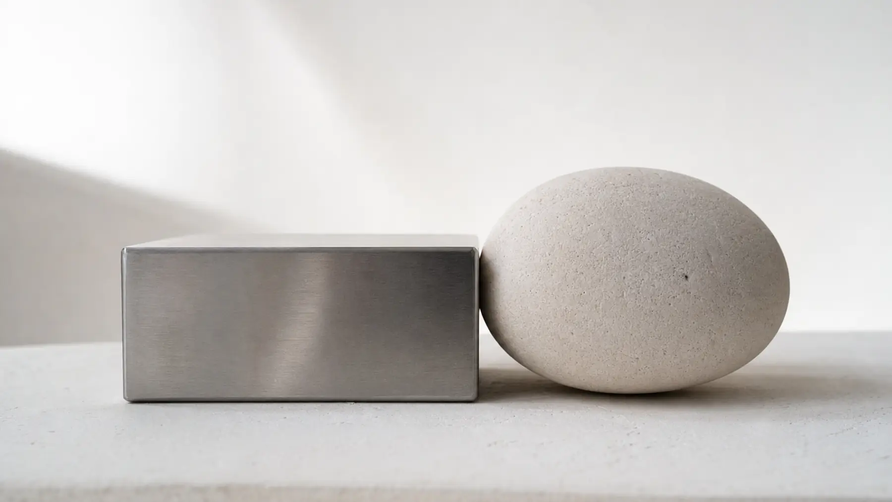 A brushed steel block and a smooth pale stone form of similar size resting side by side on a light surface